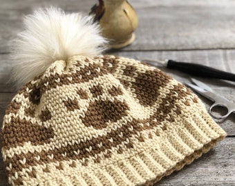 May include: A handmade knit hat in cream and brown with a faux fur pom-pom. The hat features a repeating pattern and a ribbed brim. A pair of scissors and a crochet hook are visible in the background.