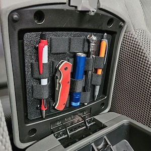 May include: Black organizer with elastic straps holding a red and white pen, a red and silver pocket knife, a blue flashlight, a silver tool, and an orange pen. The organizer is inside a car's center console.