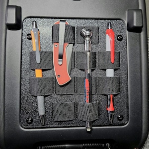 May include: Black organizer panel with elastic straps holding a red and black pocket knife, a tire pressure gauge, and two pens. The pens are orange and gray, and red and white. The panel is mounted on a black surface.
