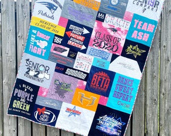 May include: A handmade quilt made from various t-shirts, featuring a patchwork design. The quilt displays a variety of colors, including pink, blue, and yellow, with text and graphics from different events and organizations. The quilt is draped over a wooden fence.