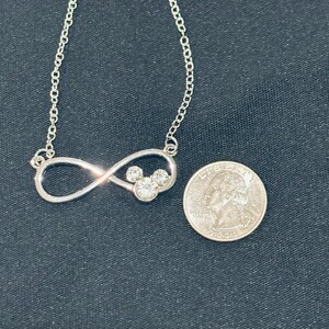 Pu&ograve; includere: Una collana color argento con un ciondolo a forma di simbolo dell'infinito che incorpora un design a testa di Topolino. Il ciondolo ha tre gemme rotonde e trasparenti. Una moneta &egrave; accanto alla collana per il confronto delle dimensioni.