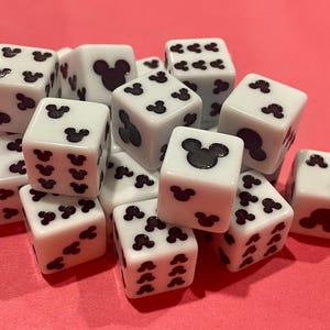 May include: A collection of white dice with black Mickey Mouse silhouettes replacing the traditional pips. The dice are scattered on a pink surface. The dice are a fun and playful item.