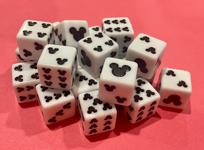 May include: A collection of white dice with black Mickey Mouse silhouettes replacing the traditional pips. The dice are scattered on a pink surface. The dice are a fun and playful item.