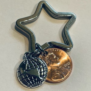 May include: A silver star-shaped keychain with a silver globe charm and a 2023 US penny attached. The penny has the words "In God We Trust" and "Liberty" on it.