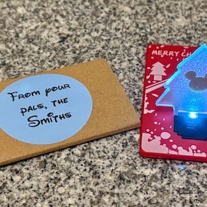 May include: A brown card with a blue circle sticker that says "From your pals, the Smiths".  A red card with a white snowflake design and a clear plastic Christmas tree with a blue light and a Mickey Mouse silhouette.