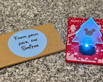 May include: A brown card with a blue circle sticker that says "From your pals, the Smiths".  A red card with a white snowflake design and a clear plastic Christmas tree with a blue light and a Mickey Mouse silhouette.