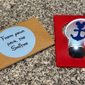 May include: A red card with a clear light-up anchor with a Mickey Mouse head on top. The card has the text "From your pals, the Smiths" on a blue circle sticker.