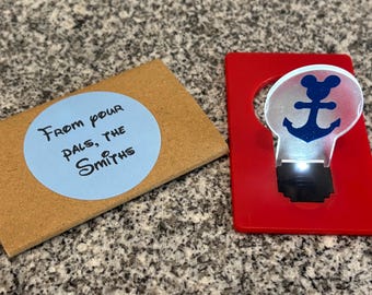 May include: A red card with a clear light-up anchor with a Mickey Mouse head on top. The card has the text "From your pals, the Smiths" on a blue circle sticker.