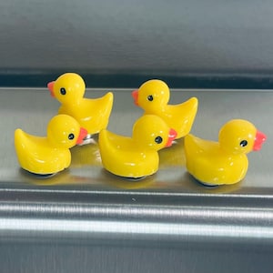 May include: Five small yellow rubber duck figurines with orange beaks. The ducks are arranged in a row on a silver surface.