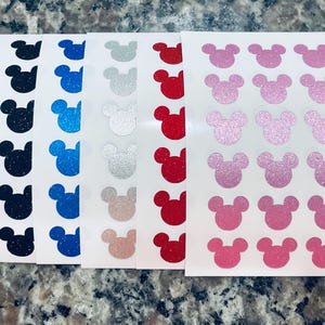 Puede incluir: Cinco hojas de papel blanco con filas de pegatinas de cabeza de Mickey Mouse con purpurina en negro, azul, plateado, rojo y rosa.