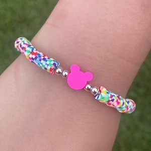May include: A colorful bracelet featuring a pink Mickey Mouse head charm and multi-colored beads. The bracelet has silver-colored accents and is set against a blurred green background.