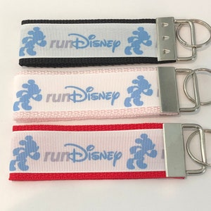 May include: Three keychains with white fabric and a blue Mickey Mouse silhouette. The fabric has the text "runDisney" printed on it. The keychains are black, pink, and red.