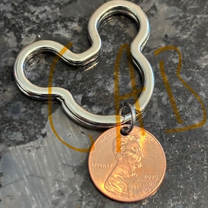 May include: Silver Mickey Mouse shaped keychain with a 2023 US penny attached. The penny has the words "In God We Trust" and "Liberty" on it.
