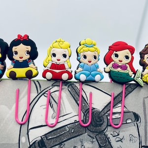 May include: Set of six colorful Disney princess character bookmark clips. Each clip features a different princess, including Jasmine, Snow White, Cinderella, Ariel, Belle, and Aurora. The clips are made of metal and have a pink hook.