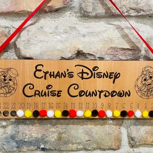 May include: Wooden sign with the text "Ethan's Disney Cruise Countdown" and a cartoon character on each side. The sign has 24 numbered circles with colorful pom-poms in some of the circles.