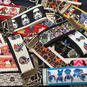 May include: A collection of fabric keychains featuring various colorful character prints, including Star Wars stormtroopers, Disney figures, and Stitch, each attached to a silver-toned metal hardware clasp and a circular split ring for keys.