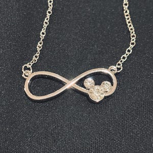 Pu&ograve; includere: Collana argentata con un ciondolo a forma di simbolo dell'infinito, che incorpora un design di Mickey Mouse con cristalli trasparenti. La delicata catena e il ciondolo risaltano su uno sfondo di tessuto scuro.