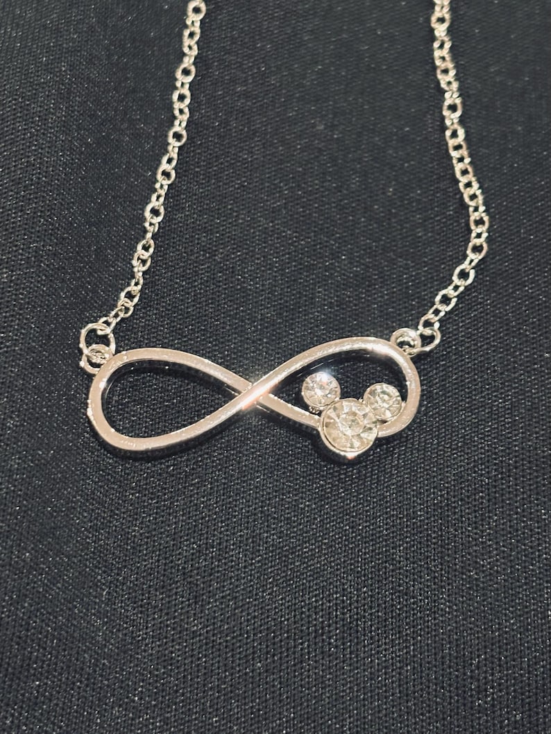Pu&ograve; includere: Collana argentata con un ciondolo a forma di simbolo dell'infinito, che incorpora un design di Mickey Mouse con cristalli trasparenti. La delicata catena e il ciondolo risaltano su uno sfondo di tessuto scuro.