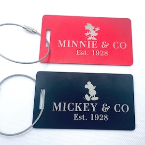 May include: Two luggage tags with black and red backgrounds. The red tag features a white silhouette of Minnie Mouse and the text "Minnie & Co Est. 1928". The black tag features a white silhouette of Mickey Mouse and the text "Mickey & Co Est. 1928".