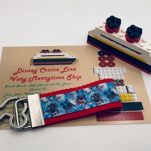 May include: A red and blue keychain with a Disney Cruise Line Very Merrytime Ship design. The keychain features a small Lego ship with red and black smoke stacks and a white deck. The text "Disney Cruise Line Very Merrytime Ship" is printed on a brown card behind the keychain.