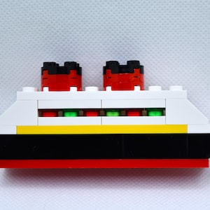 May include: A white, red, black, and yellow toy ship with green windows. The ship has two red and black smokestacks.