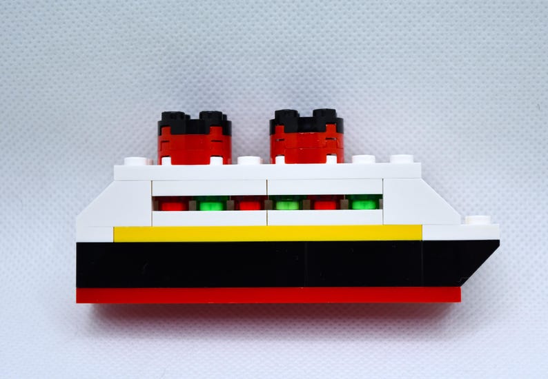 May include: A white, red, black, and yellow toy ship with green windows. The ship has two red and black smokestacks.