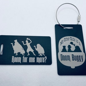 May include: Two black luggage tags with silver text and images. The first tag says "Room for one more?" and features a silhouette of the ghosts from the Haunted Mansion ride. The second tag says "My other ride is a Doom Buggy" and features a silhouette of the ghosts from the Haunted Mansion ride in a Doom Buggy.