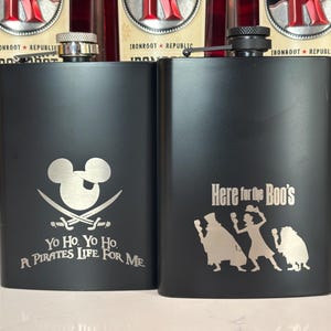 May include: Two black flasks with silver tops. One flask features a Mickey Mouse head with crossed swords and the text "Yo Ho. Yo Ho. A Pirates Life For Me." The other flask has the text "Here for the Boo's" with a silhouette of three figures.