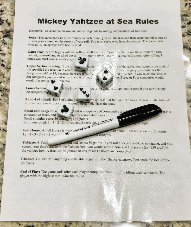 May include: A sheet of paper with the rules for "Mickey Yahtzee at Sea" is shown, along with five dice and a black dry-erase marker. The dice have Mickey Mouse icons and numbers.