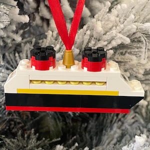 May include: A Lego ornament of a red, white, and black ship with a gold top. The ship is hanging from a red ribbon on a snowy Christmas tree.