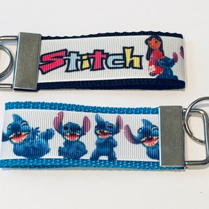 May include: Two Stitch keychains. The first keychain is black with white text that says "Stitch" and a picture of Stitch and Angel. The second keychain is blue with white stripes and multiple pictures of Stitch.