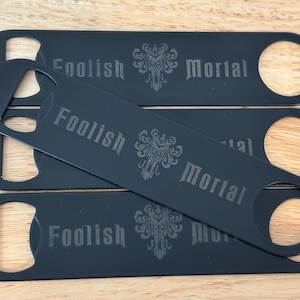 May include: Four black bottle openers with the words "Foolish Mortal" and a decorative emblem etched on each. The openers have a rounded end with a circular opening for leverage and a flat, rectangular body.