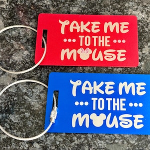 May include: Two luggage tags with silver keychains. The red tag says "Take me... to the... mouse" with a pink mouse ear silhouette. The blue tag says "Take me... to the... mouse" with a white mouse ear silhouette.