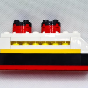 May include: A white, red, black, and yellow Lego model of a cruise ship with two red smokestacks.