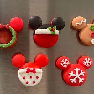 May include: Five Christmas-themed Mickey Mouse magnets. The magnets are red, white, green, and brown. One magnet features a wreath, one has a Santa hat, one has a gingerbread man, and two have snowflakes.