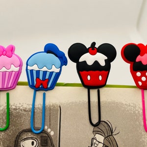 May include: Four colorful cupcake-shaped bookmark clips. The clips are made of plastic and feature different cartoon characters. The clips are attached to metal bookmark holders.