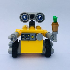 May include: A yellow Lego robot with gray arms and wheels, holding a small brown container with green leaves. The robot has large, blue-eyed optics and a control panel on its front.