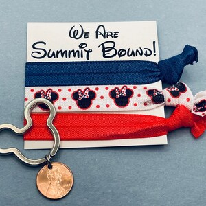 May include: Three red, white, and blue hair ties with a Minnie Mouse design. The hair ties are tied together with a blue hair tie. The hair ties are on a white card with the text "We Are Summit Bound!" The hair ties are next to a silver Mickey Mouse shaped keychain with a penny attached.