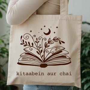 May include: Beige tote bag with a brown illustration of an open book, leaves, a crescent moon, and stars. The text "kitaabein aur chai" is printed below the book design. The bag has two handles.