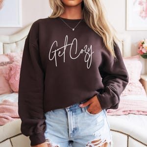 May include: A dark brown sweatshirt with the words "Get Cozy" in white script. The long-sleeved top is paired with blue denim shorts. The model is wearing a silver necklace.