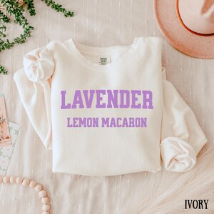May include: Ivory-colored sweatshirt with the words "LAVENDER" and "LEMON MACARON" printed in purple. The sweatshirt is made of a soft material and is folded on a white surface. A hat is visible in the background.