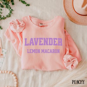 May include: A light pink sweatshirt with the words "LAVENDER" and "LEMON MACARON" printed in purple block letters. The word "PEACHY" is printed in the bottom right corner. The sweatshirt is folded on a textured surface.