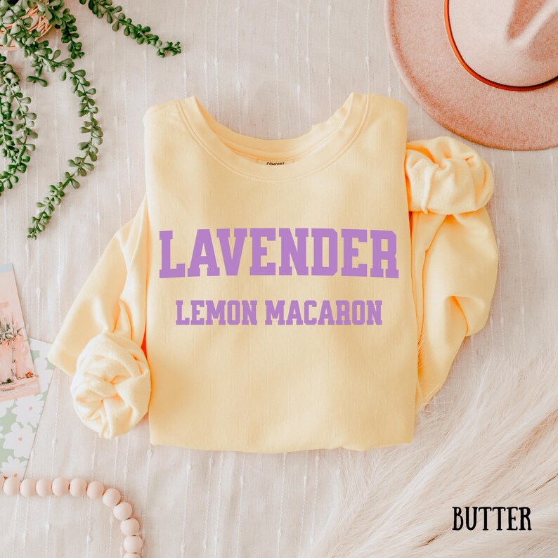 May include: A butter-yellow sweatshirt with the words "LAVENDER" and "LEMON MACARON" printed in lavender. The word "BUTTER" is printed in the bottom right corner. The long-sleeved top is displayed on a white surface.