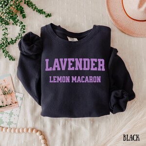 May include: Black crewneck sweatshirt with the text "LAVENDER LEMON MACARON" in purple block letters. The word "BLACK" is printed in the bottom right corner. The sweatshirt is displayed on a white surface.