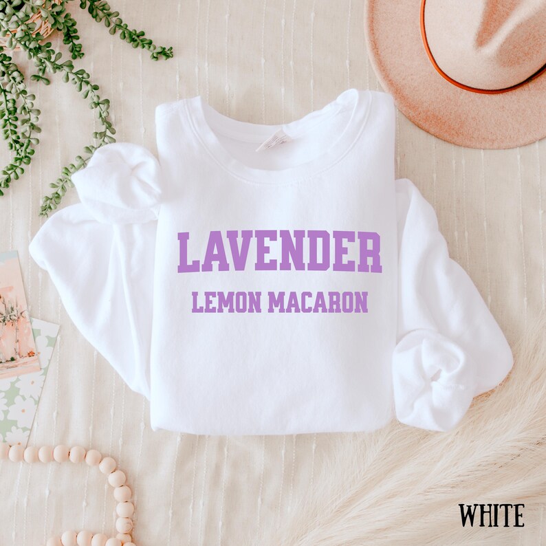 May include: A white sweatshirt with the words "LAVENDER" and "LEMON MACARON" printed in purple. The word "WHITE" is printed in the bottom right corner. The sweatshirt is made of a soft material.