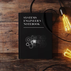 May include: A black notebook with the title "Systems Engineer's Notebook" on the cover. The cover also features a design of gears and a circuit board. The notebook is on a wooden surface with two Edison bulb lights hanging above it.