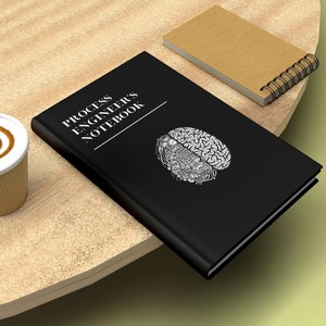 May include: A black notebook with the text "Process Engineers Notebook" and a stylized brain graphic. The notebook is on a wooden table with a cup of coffee and a brown spiral notebook.