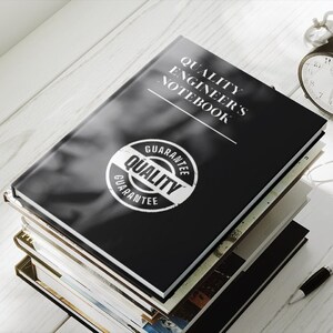 May include: A black notebook with the text "Quality Engineer's Notebook" and a white circle with the text "Guarantee Quality" on the cover. The notebook is stacked on top of other books on a white wooden surface.