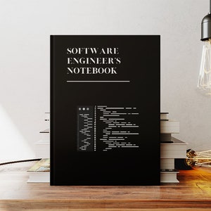 Puede incluir: Una libreta negra con el texto "SOFTWARE ENGINEER'S NOTEBOOK" en la portada. La libreta est&aacute; sobre una mesa de madera con una pila de libros detr&aacute;s.