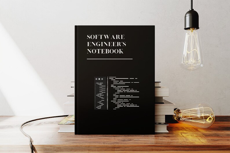 Puede incluir: Una libreta negra con el texto "SOFTWARE ENGINEER'S NOTEBOOK" en la portada. La libreta est&aacute; sobre una mesa de madera con una pila de libros detr&aacute;s.